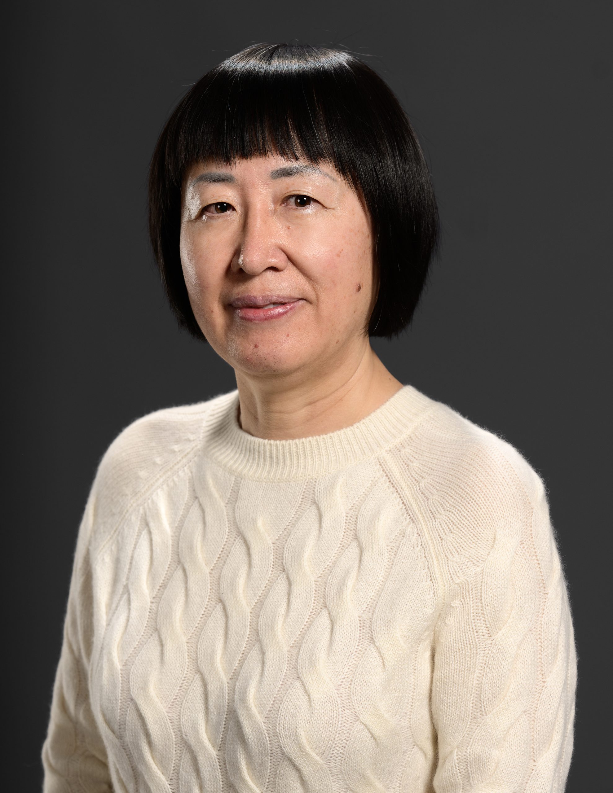Professional studio portrait of a middle-aged Asian woman wearing a cream cable-knit sweater against a gray background.