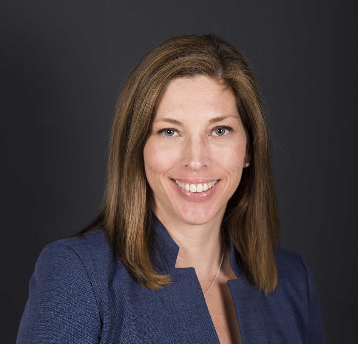 Professional headshot of a smiling woman with shoulder-length brown hair wearing a dark blue blazer over a white top.
